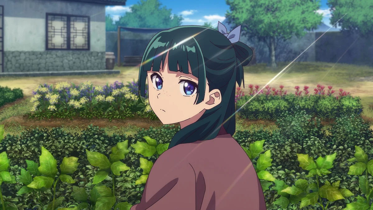 Maomao turns toward the viewer among green plants, with tiled buildings behind her.