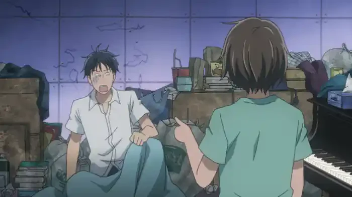 Chiaki reacts to Nodame in a room crowded with bags and containers beside a piano.