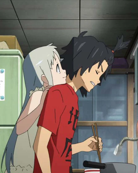 Menma stands close behind Jinta while he holds chopsticks in a kitchen.
