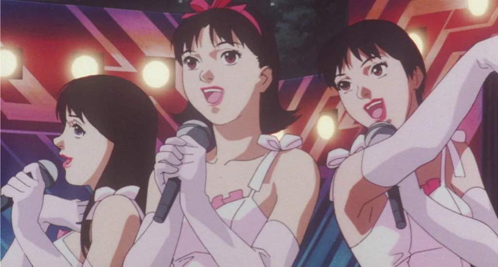 Three members of CHAM! sing into microphones in coordinated pink and white stage outfits beneath bright lights.