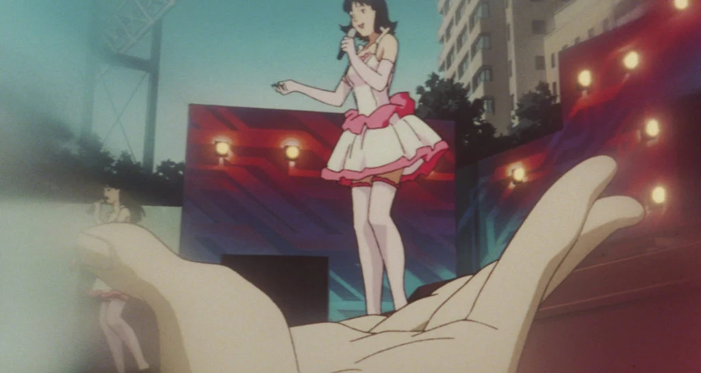 Mima performs on a brightly lit stage while a large outstretched hand in the foreground appears to hold her.