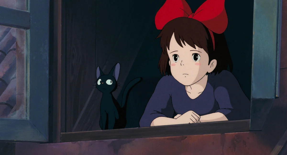 Kiki leans on a wooden window ledge with her hands folded while Jiji stands beside her.