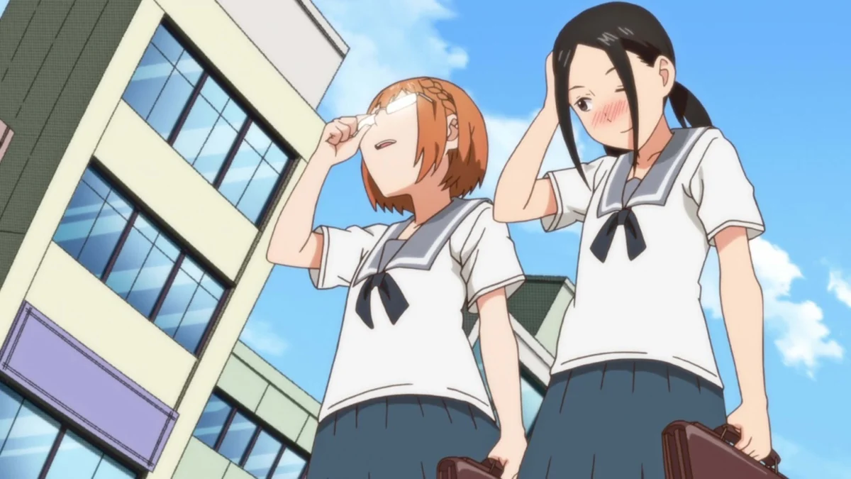 Two schoolgirls stand beside city buildings; the orange-haired girl touches her glasses while her dark-haired companion blushes.