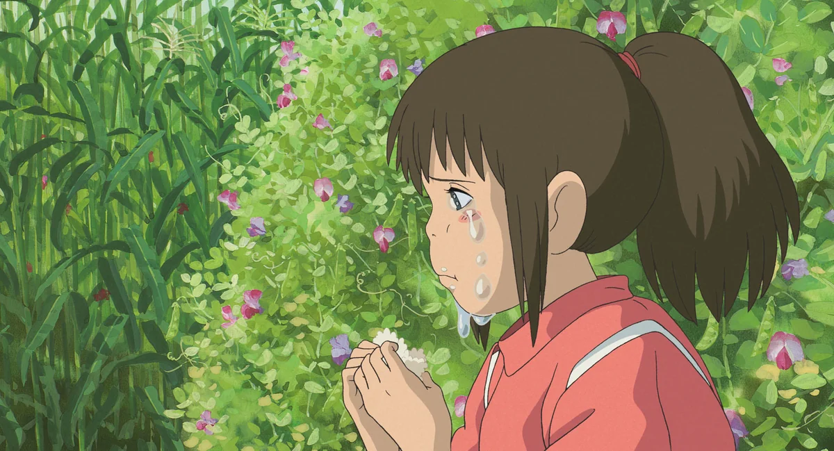 Chihiro holds a rice ball near her mouth, with tears on her cheeks and a flowering hedge behind her.