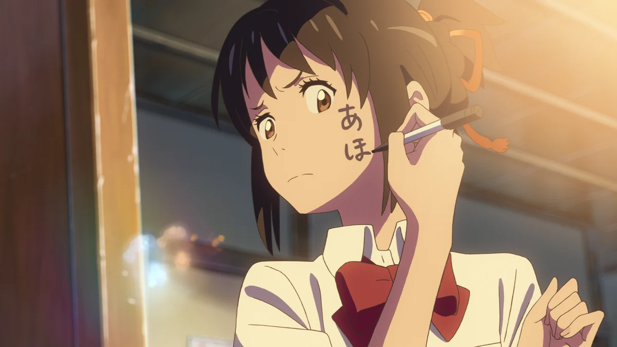 Mitsuha's face appears in a mirror, with a marker held beside characters written on her cheek.