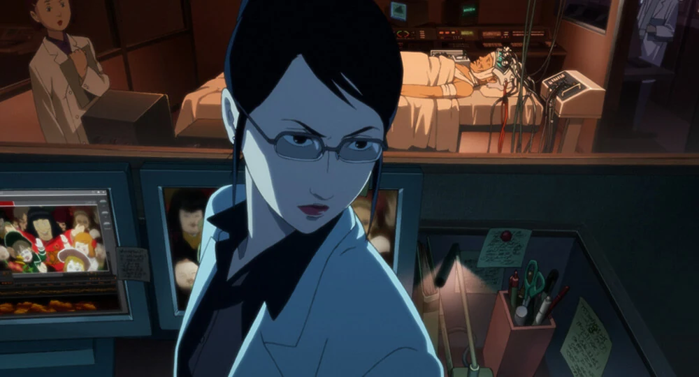 Atsuko Chiba stands before monitors, with a patient and medical equipment visible through the window behind her.