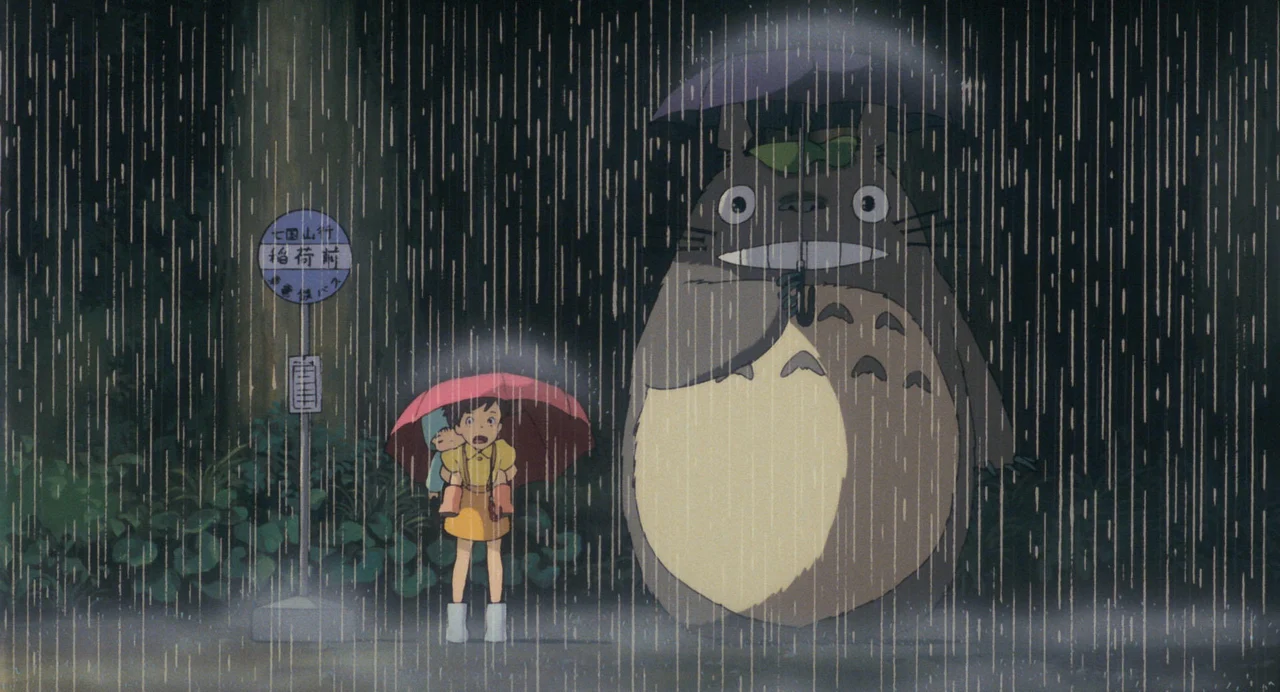 Satsuki carries Mei beside Totoro beneath umbrellas in the rain.