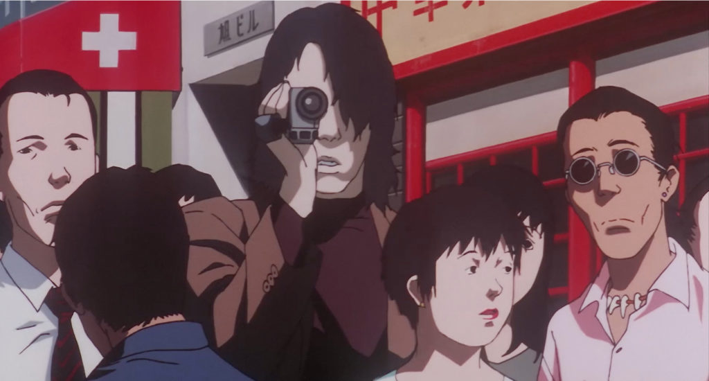 A dark-haired man points a video camera towards the viewer from a crowd outside shops.