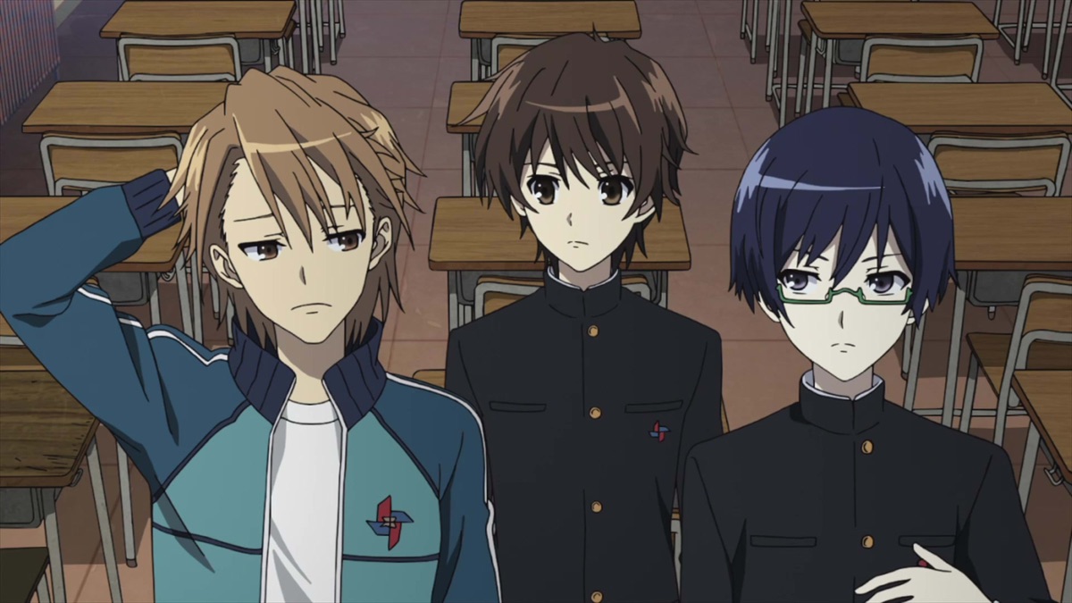 Three boys stand in a classroom with empty desks behind them; one scratches the back of his head.