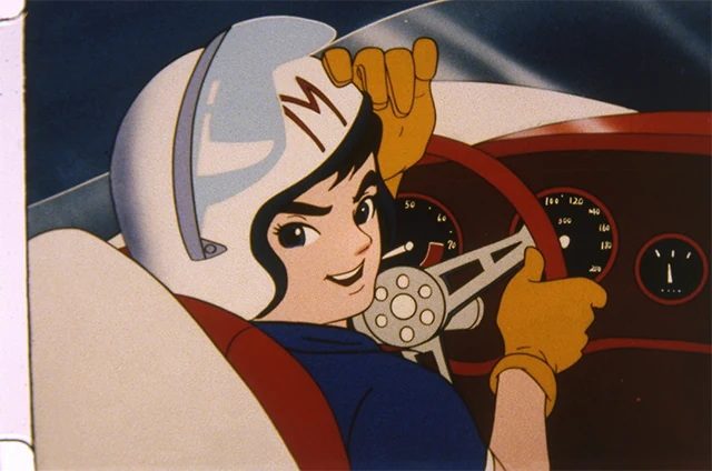 A helmeted driver turns towards us from the red cockpit of the white Mach 5.