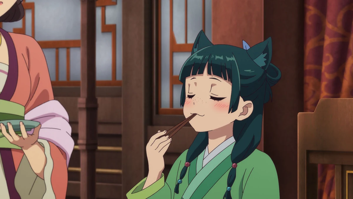 Maomao holds chopsticks to her mouth, with closed eyes and cartoon cat ears, as another attendant holds a dish.